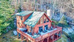 A log cabin with a porch and deck, surrounded by trees and a forested area. - 3 Bedroom, Beautiful Mountain Views, Wireless Internet, Located behind MVCC (MAGGIE VALLEY)