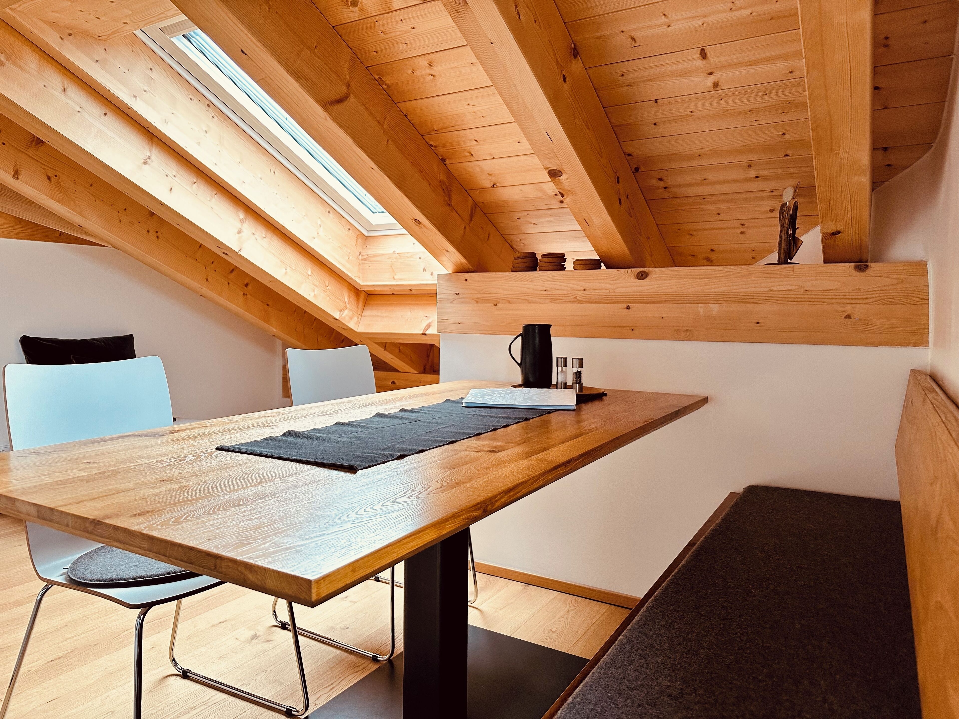 A modern loft bedroom with a sloped ceiling, skylight, wooden beams, a wooden table, a chair, and a bench.