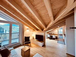 A spacious living area with wooden beams, a large sliding glass door, a cozy seating area, and a kitchen in the background. - Alpenglück-Garmisch, new top-floor vacation apartment (Garmisch-Partenkirchen)