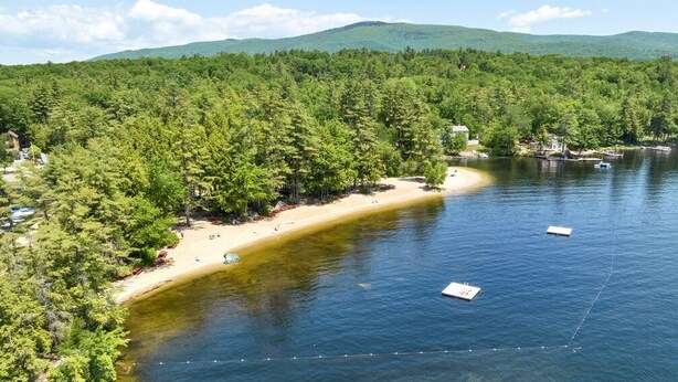 Propiedad frente a un lago con playa de arena, bosque frondoso y barcos.