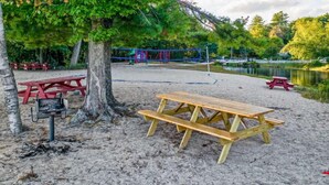 Zona de playa arenosa con mesas de picnic y un parque infantil al fondo.