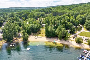 An aerial view of a lakeside property with a dock, a sandy beach, and a parking area surrounded by dense forest.