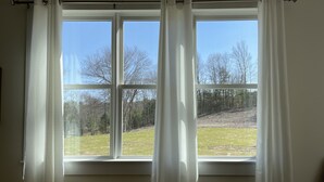 A window with white curtains, a view of a grassy area with trees, and a clear sky. - Magnificent Modern Farmhouse (Mountain Dale)