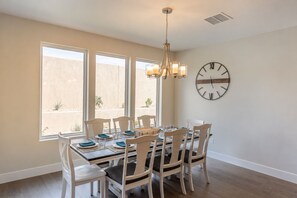 A dining area with a table set for six, white chairs, a chandelier, and a wall clock. - Golfside 4BR - 3 King Suites, w/ Hot Tub & Firepit (St. George)