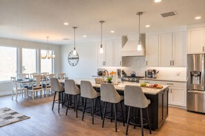 A modern kitchen with a central island, bar stools, and a dining area with chairs. - Golfside 4BR - 3 King Suites, w/ Hot Tub & Firepit (St. George)