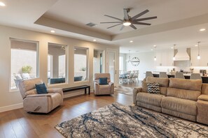 A spacious living room with a large sofa, a recliner, and a coffee table. There is a dining area with a chandelier and a kitchen in the background. - Golfside 4BR - 3 King Suites, w/ Hot Tub & Firepit (St. George)