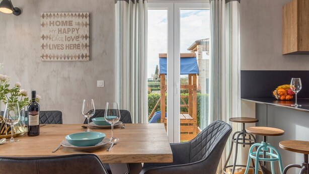 Un comedor con una mesa de madera, sillas y una botella de vino. Se ve un balcón con un toldo azul y una terraza de madera.