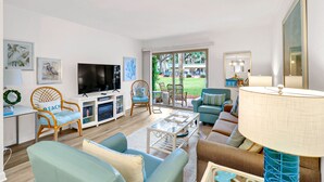 A spacious living room with a flat-screen TV, comfortable seating, a glass coffee table, and a view of a garden through large sliding doors. - Pet Friendly Beachside Villa - Across the Street from the Beach! (Hilton Head Island)