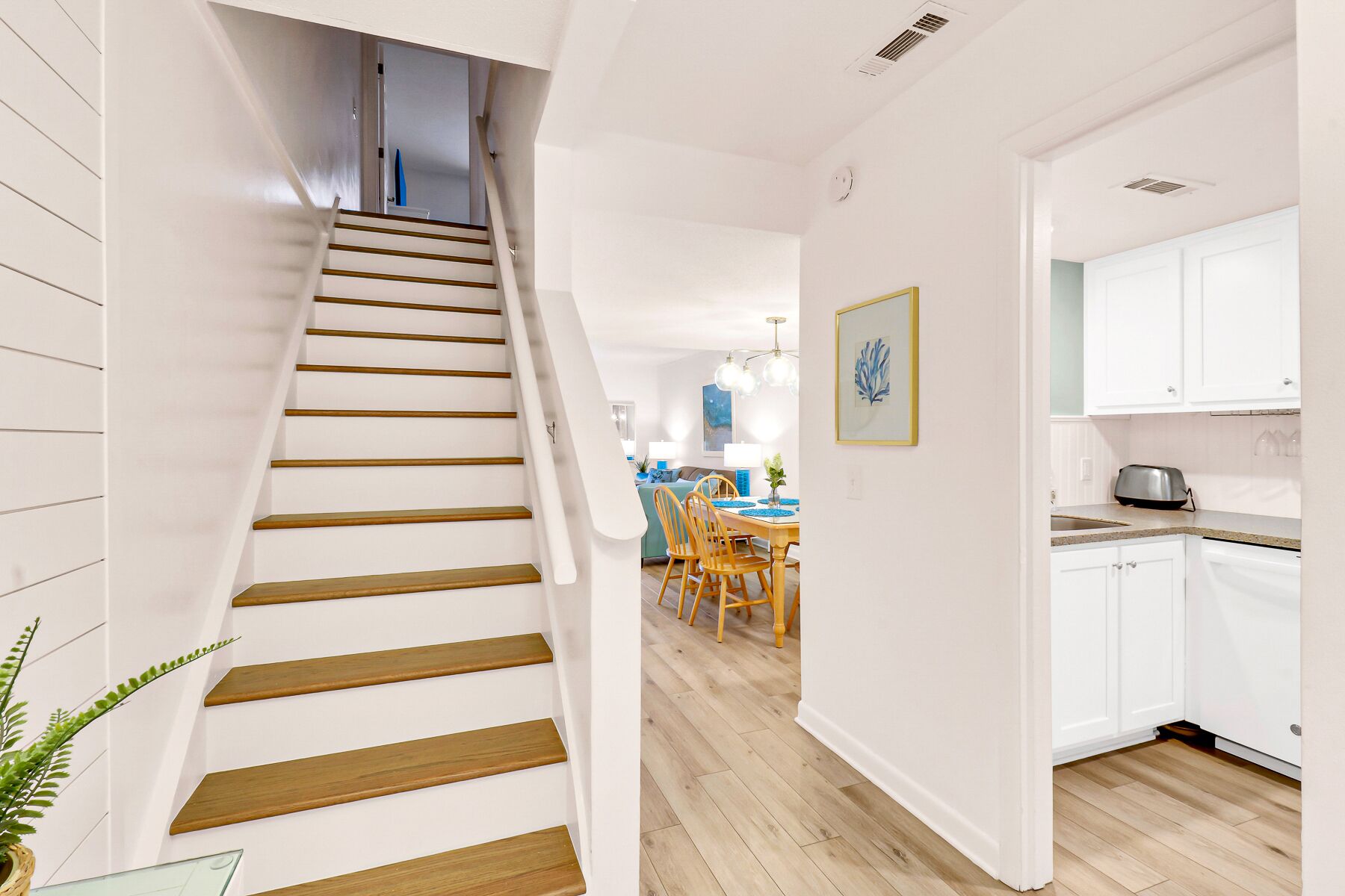 Un escalier mène à une salle à manger dotée d’un plancher en bois, de murs blancs et d’une cuisine avec des meubles blancs.