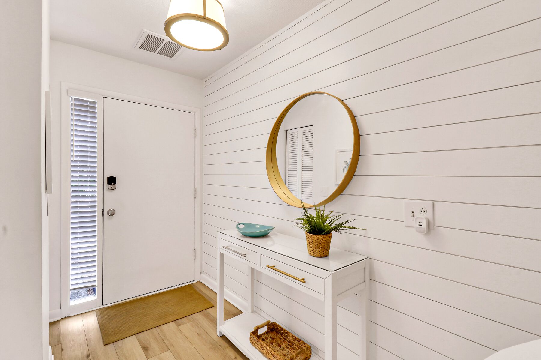 A hallway with white walls, a white door, a white console table with a mirror, a bowl, and a plant.