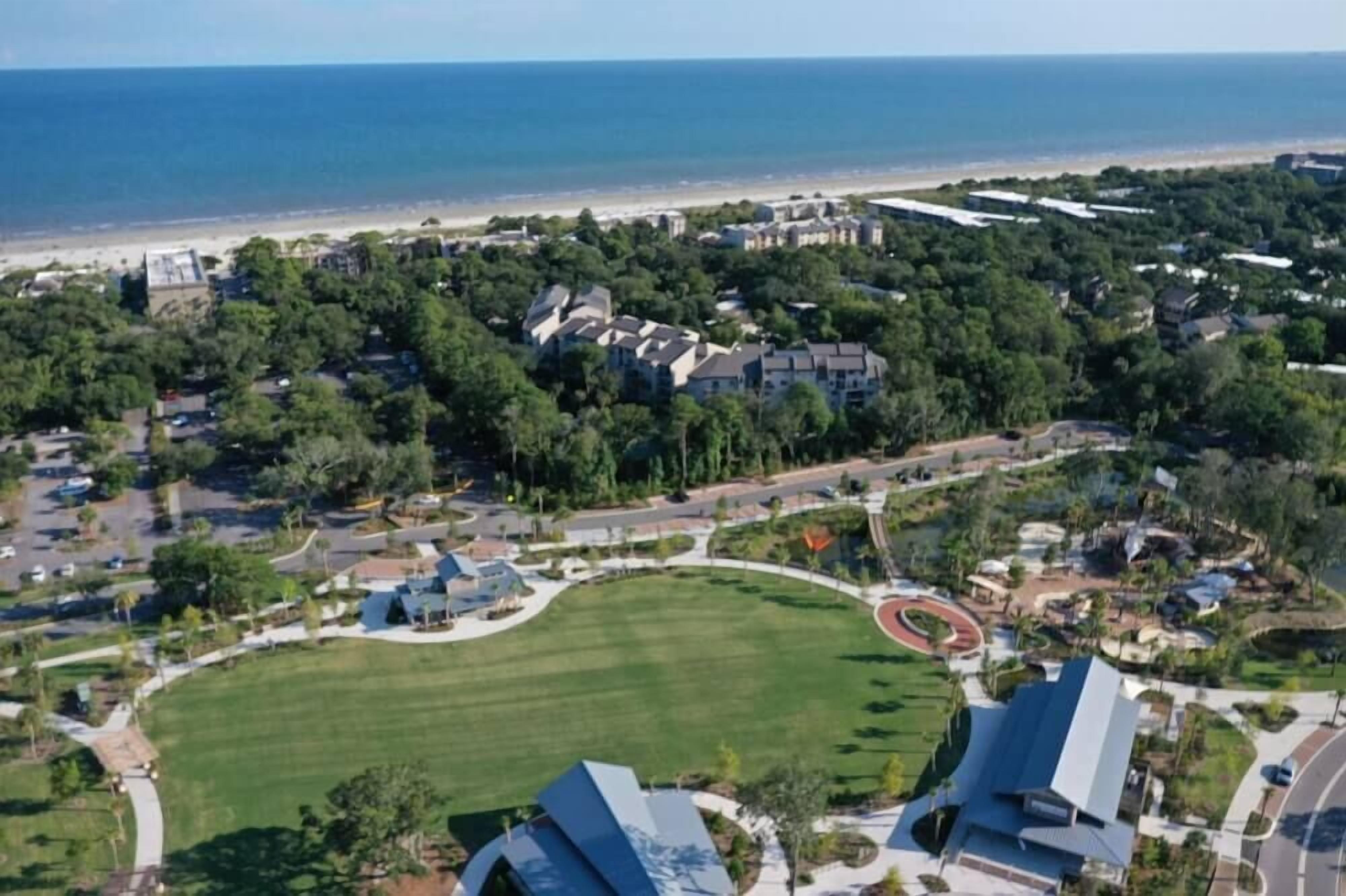 Vue aérienne d’un quartier résidentiel situé sur la côte, avec des maisons, un grand parc verdoyant et une plage en arrière-plan.