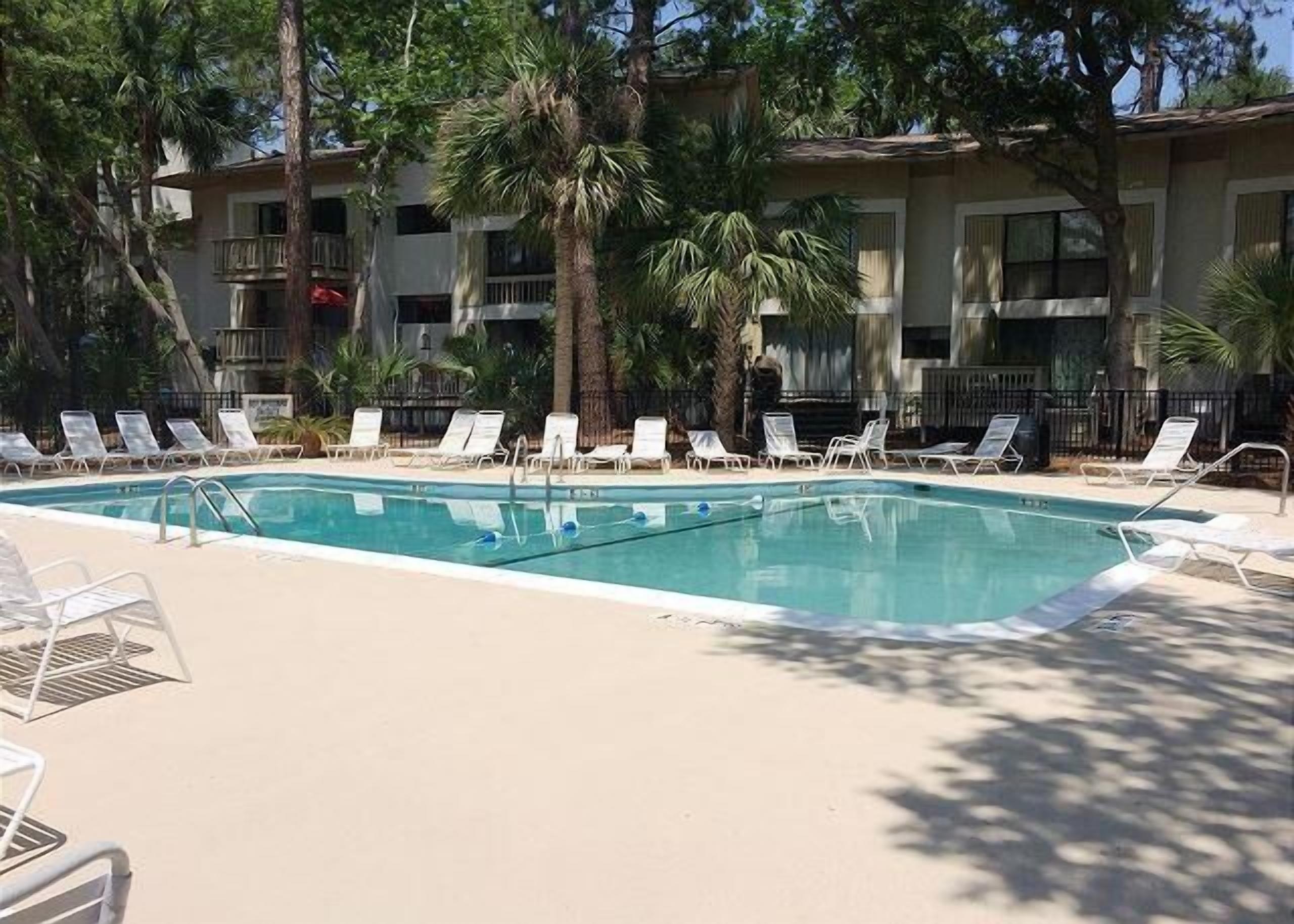 A rectangular swimming pool with a clear blue water surface, surrounded by white lounge chairs and palm trees.