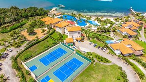 An aerial view of a resort with multiple buildings, a large swimming pool, tennis courts, and a beachfront. - Pristine Bay Villa 1304 with large pool - 3 bedroom 3 Condo by RedAwning (Roatan)