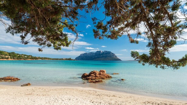 Een strand met helder turquoise water, een rotsachtige landtong en een berg op de achtergrond.