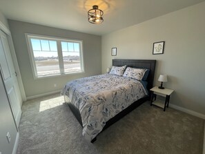Une chambre avec une grande fenêtre, un lit recouvert d’une literie à motifs floraux, une table de chevet avec une lampe et un luminaire au plafond.