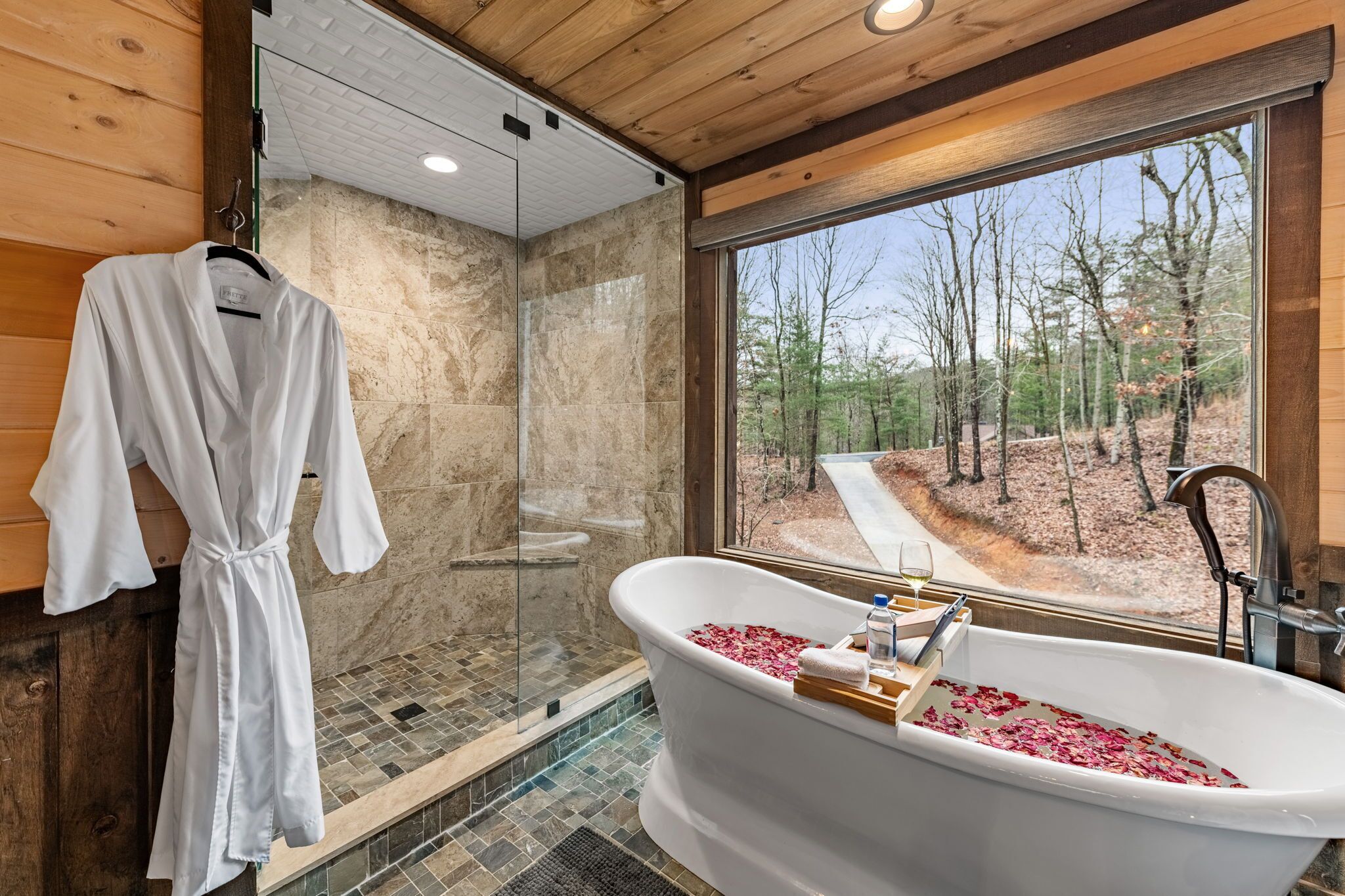 Un bagno con una vasca freestanding, un box doccia in vetro e vista su una foresta attraverso un'ampia finestra.