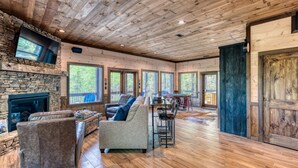 A spacious living room with a stone fireplace, wooden ceiling, and large windows offering views of the outdoors. - Luxury Mountain Cabin for Families & Groups | 4 King Bedrooms + Hot Tub (Epworth)