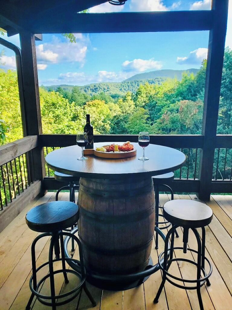 A wooden deck with a round table and two stools, a bottle of wine, and a plate of food.