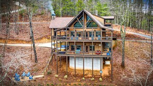 A multi-story log cabin with a central deck, surrounded by a forest. - Luxury Mountain Cabin for Families & Groups | 4 King Bedrooms + Hot Tub (Epworth)