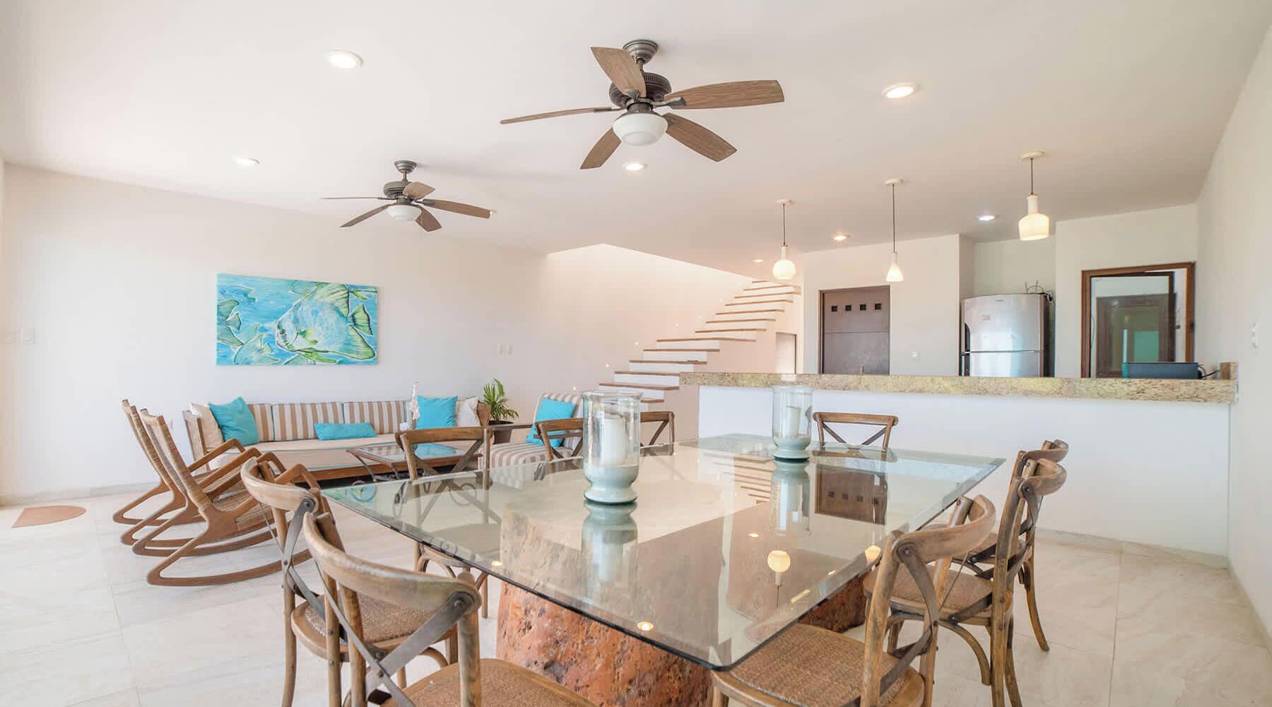A modern kitchen with a built-in refrigerator, a dining area with a glass table and wooden chairs, and a staircase leading to an upper level.