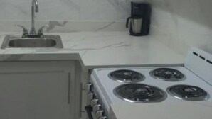 Una cocina compacta con gabinetes blancos, estufa, pileta y ventana con persianas.