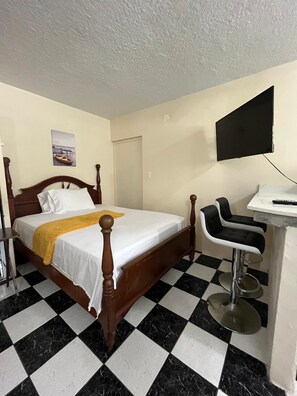 Habitación de hotel con piso a cuadros, cama con cabecera de madera, escritorio con silla y televisor de pantalla plana montado en la pared.