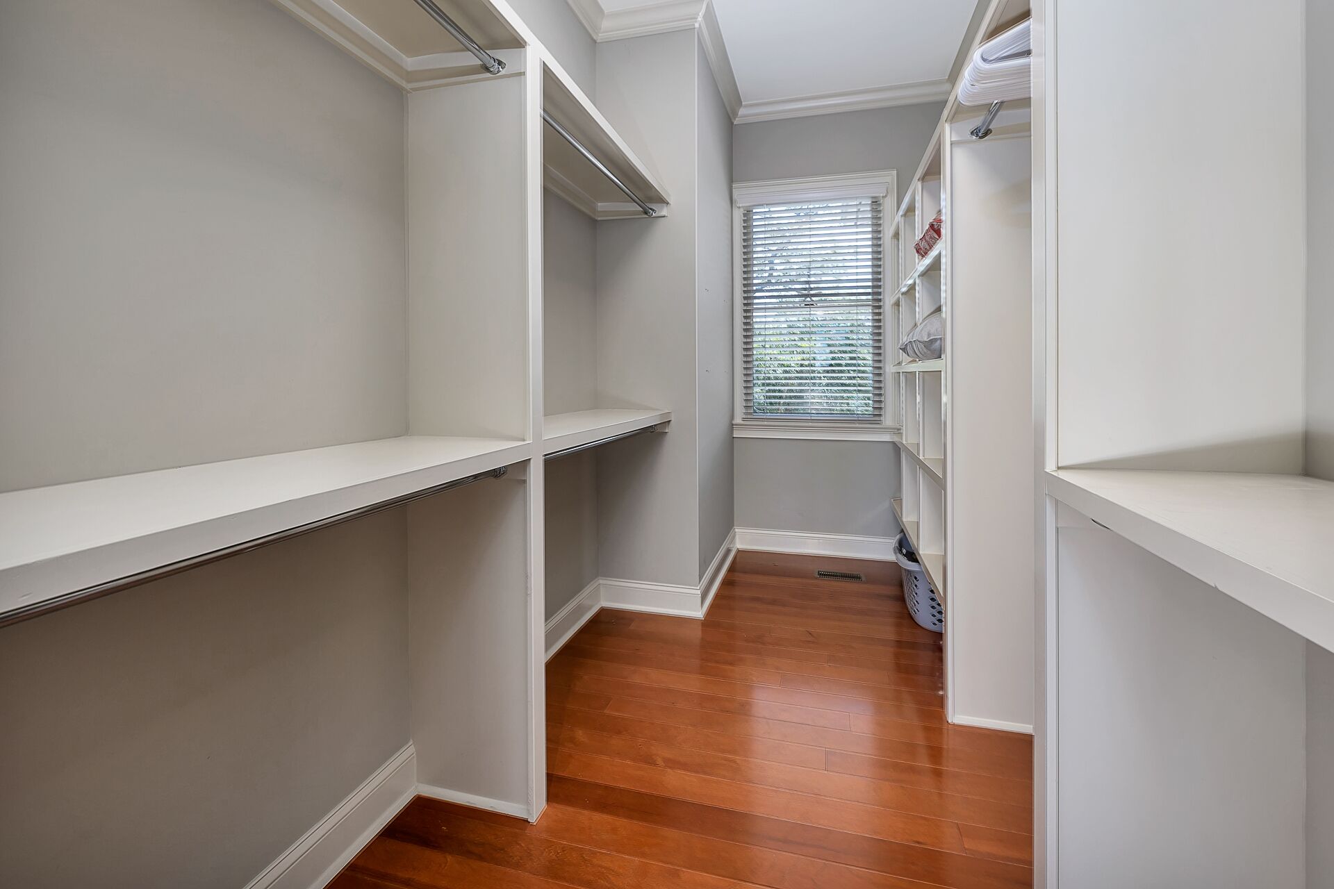 A narrow walk-through closet with white shelving and a window with blinds.