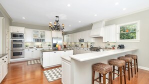 Una cocina moderna con armarios blancos, una isla central con taburetes de bar, electrodomésticos de acero inoxidable y una lámpara de araña.