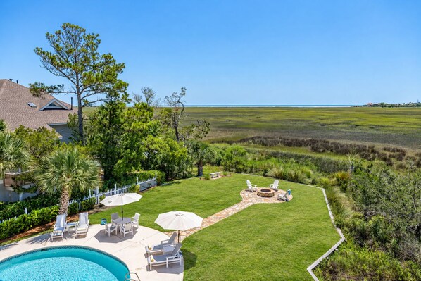 A backyard with a swimming pool, lounge chairs, a fire pit, and a view of the ocean. - High Tide - BREATHTAKING Marsh and Ocean Views! Private Pool, Fire Pit (Saint Simons Island)