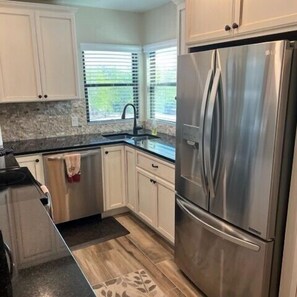 Una cocina con armarios blancos, un refrigerador de acero inoxidable, un ventanal con persianas y una alfombra estampada.