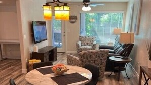 A modern living room with a round wooden dining table, black chairs, a patterned armchair, a sofa, and a television.