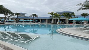 Une grande piscine entourée de palmiers et de bâtiments modernes, avec des chaises longues et des parasols à proximité.