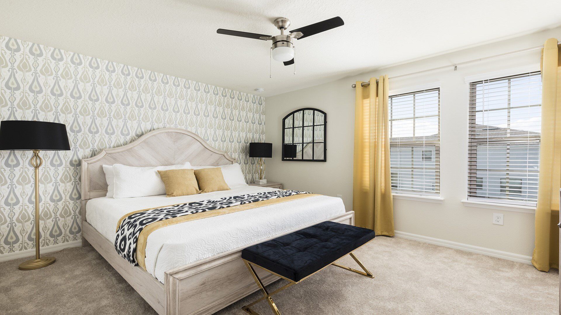 A bedroom with a bed, a ceiling fan, a nightstand, a lamp, a window with blinds, and a wall-mounted mirror.