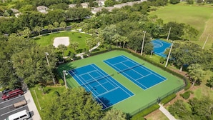 Vista aerea di un campo da tennis circondato da vegetazione lussureggiante e edifici residenziali.