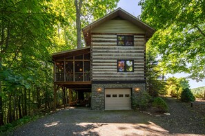 A log cabin with a stone foundation, a garage, and a deck overlooking a forested area. - Rustic Log Cabin Near Todd with Hot Tub, Fire Pit & Mountain Views (Todd)
