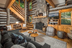 A log cabin interior with a stone fireplace, wooden staircase, and a cozy seating area. - Rustic Log Cabin Near Todd with Hot Tub, Fire Pit & Mountain Views (Todd)