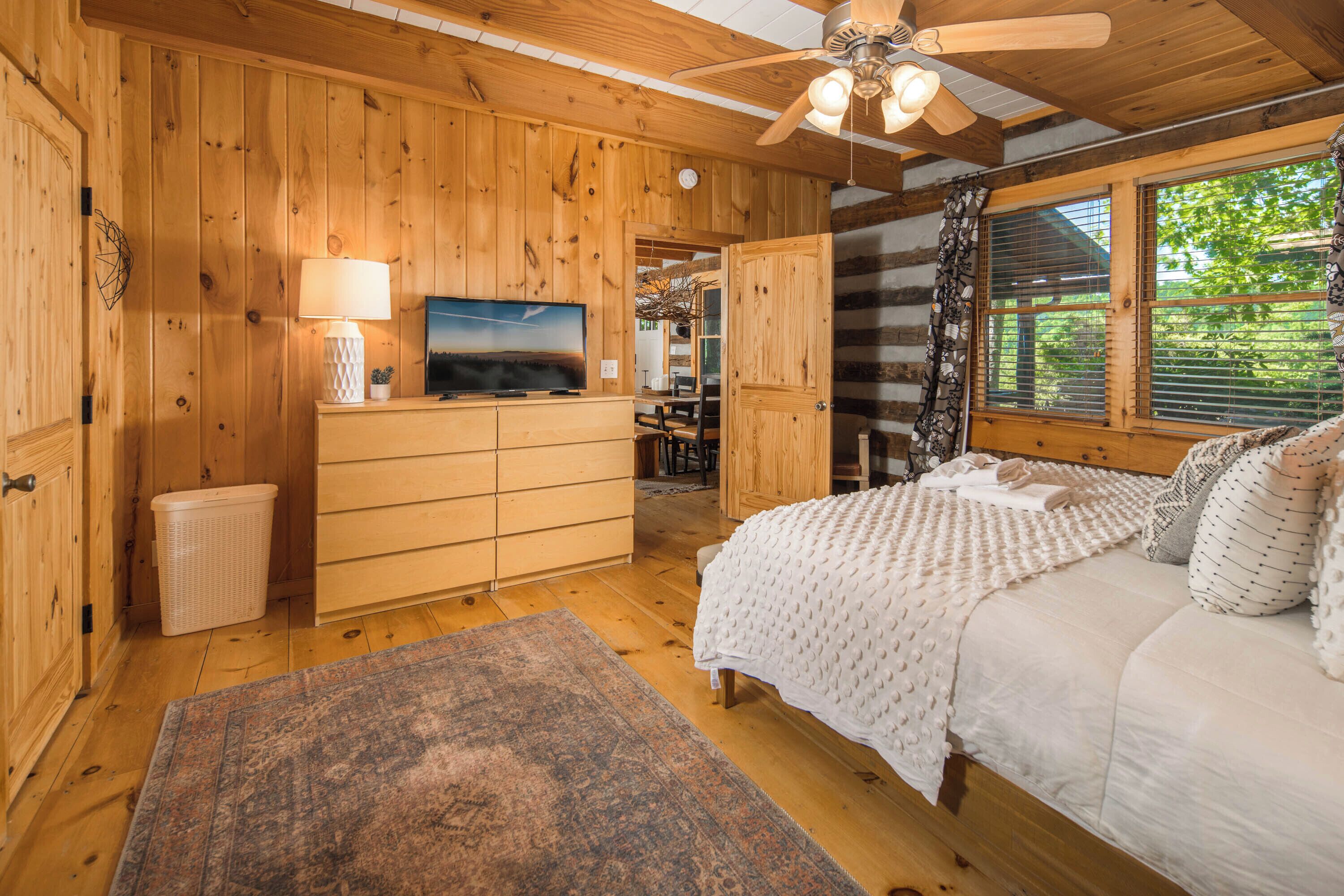 A wooden cabin bedroom with a bed, a TV, a ceiling fan, and a window with curtains.
