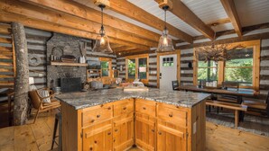 A log cabin interior with a central island kitchen, wooden cabinets, and a stone fireplace.