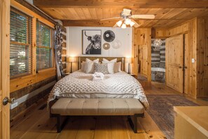 A wooden cabin bedroom with a bed, a bench, a ceiling fan, and a stone fireplace.