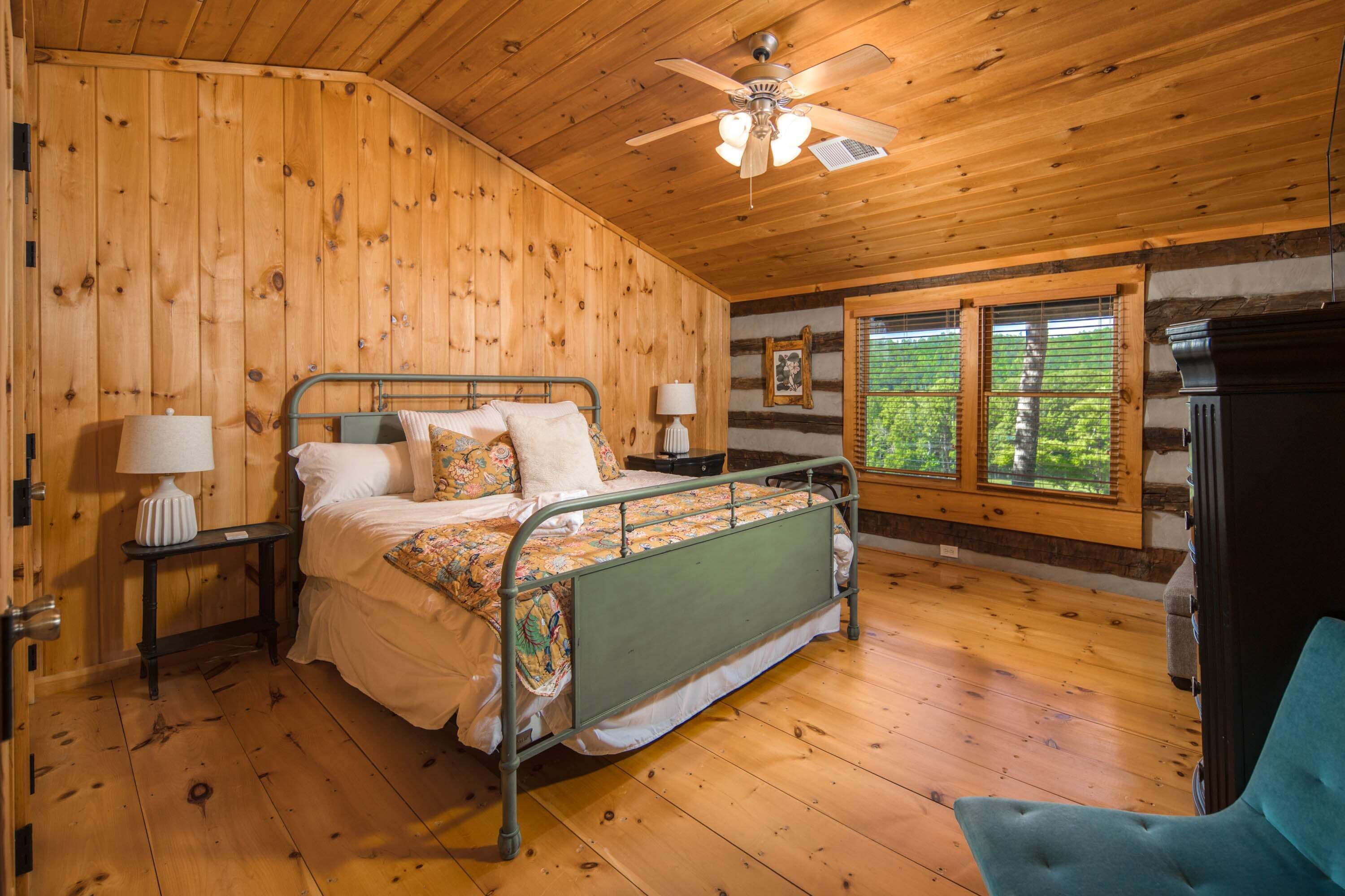 Ein Schlafzimmer mit Holzwandverkleidung, einem Metallbettgestell, zwei Nachttischen mit Lampen, einem Deckenventilator und einem großen Fenster mit Jalousien.