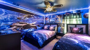 Un dormitorio con diseño temático espacial, que incluye una cama con estampado galáctico, un ventilador de techo y un mural de una nave espacial.