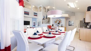 Uma sala de jantar moderna com mobiliário branco, jogos americanos vermelhos e um lustre central.