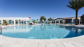 Una piscina amplia y cristalina con sillones de descanso y sombrillas, rodeada de palmeras y un pabellón blanco.