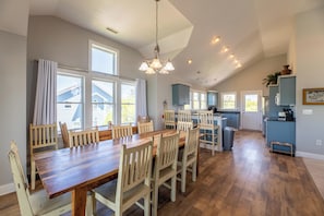Un grand espace à manger avec une table et des chaises en bois, une cuisine aux armoires bleues et une vue sur l’extérieur.
