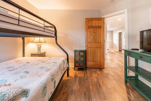 A bedroom with a bunk bed, a bedside table with a lamp, a wooden door, and a TV on a green stand. - Allora - Corolla, Ocean Sands, 450 Yds to Beach, Pool, Hot Tub, Pool Table, Gas Fireplace, Pets OK (Corolla)