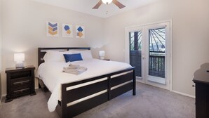 Un dormitorio con cama, mesitas de noche, ventilador de techo, una obra de arte en la pared y una puerta corrediza de vidrio al exterior.
