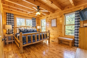 Ein Schlafzimmer in einer Holzhütte mit einem Bett, zwei Nachttischen, einem Deckenventilator und großen Fenstern.