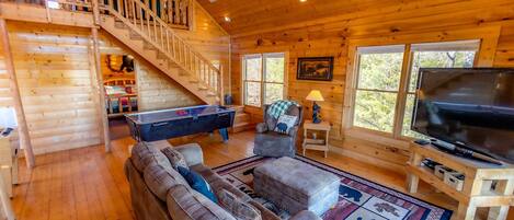 Ein gemütliches Blockhaus-Interieur mit einer Holztreppe, einem Billardtisch, einer Couch, einem Fernseher und großen Fenstern.