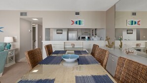 Une cuisine moderne avec des meubles blancs, un îlot central et un coin repas comprenant une table en verre et des chaises en osier.