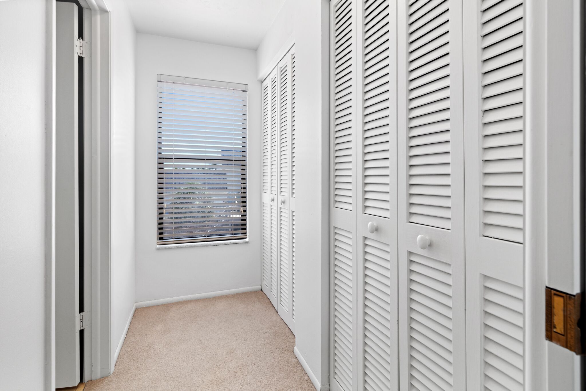 A narrow hallway with white walls, a window with blinds, and a door with louvered shutters.